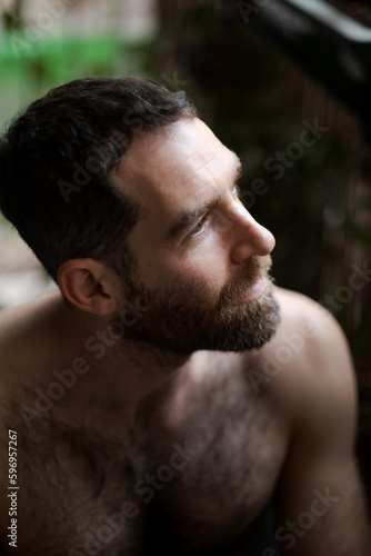 Looking over his shoulder while relaxing at home, a man expresses thoughtfulness and contemplation. He is shirtless, with a thick beard that draws attention to his face.