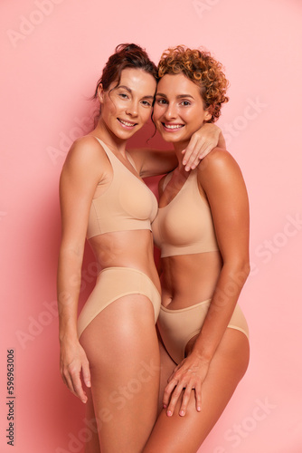 Two beautifully built girls in lingerie pose against a pink wall, dressed in a comfortable top and panties for sports, one hugging the other around the neck, friendship concept, copy space, high