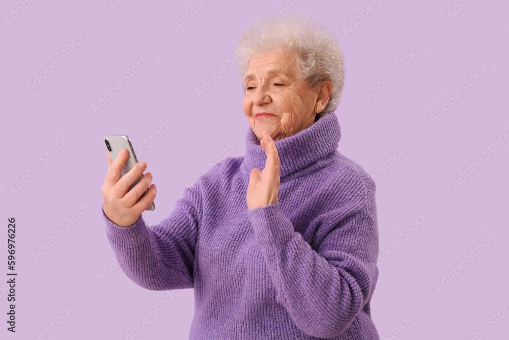 Senior woman with mobile phone video chatting on lilac background Senior woman with mobile phone video chatting on lilac background
