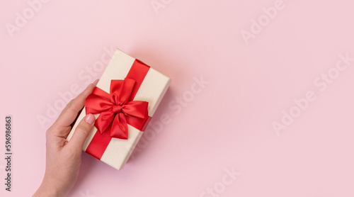 Hands holding gift box with red bow on pink background. Top view