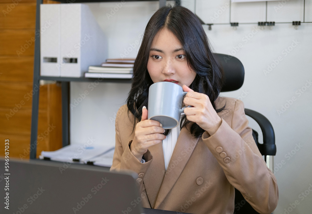 Young female worker using laptop in office, Officer holding coffee/tea mug while looking at open laptop computer on table.Cozy office workplace, online work.Businesswoman drinking coffee Young female worker using laptop in office, Officer holding coffee/tea mug while looking at open laptop computer on table.Cozy office workplace, online work.Businesswoman drinking coffee
