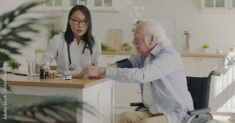 Nurse examining disabled senior man in wheelchair talking discussing treatment in apartment in kitchen. Medical care and communication with patient concept. Nurse examining disabled senior man in wheelchair talking discussing treatment in apartment in kitchen. Medical care and communication with patient concept.