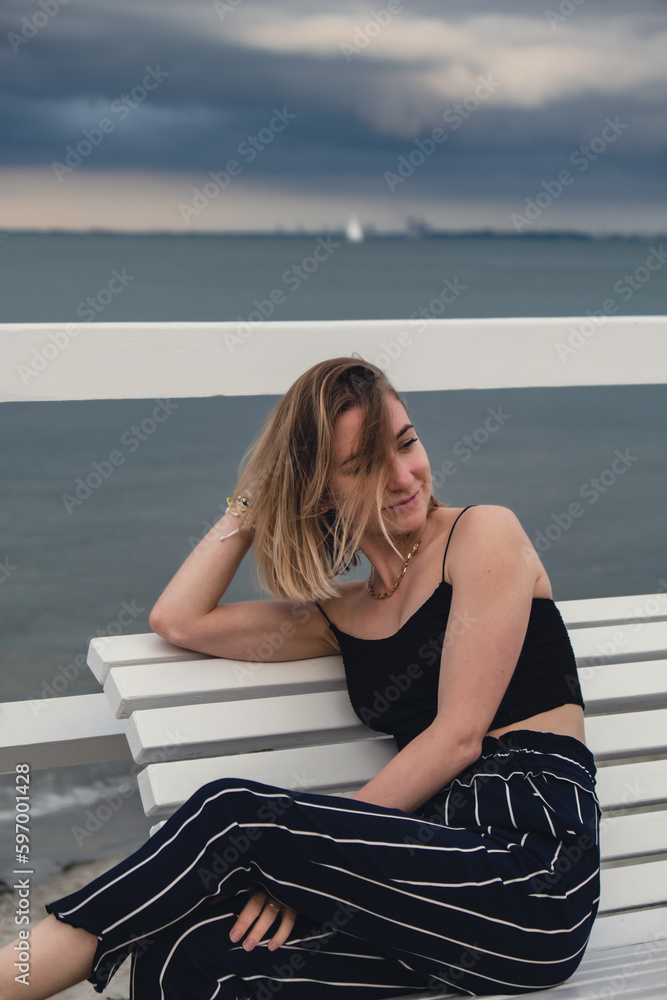 Young woman sitting on bench on wooden pier blurred beachside background. Attractive female enjoying the sea shore travel and active lifestyle concept. Springtime. Wellness wellbeing mental health