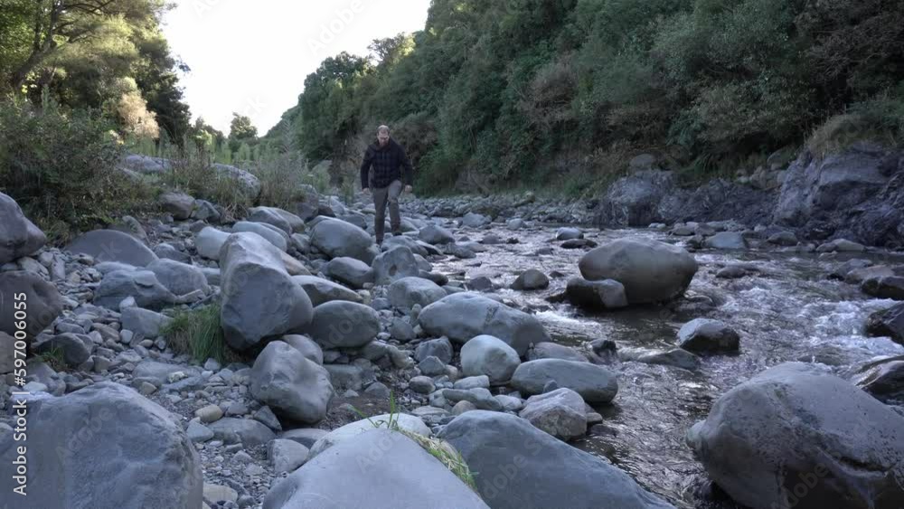 Wide shot of hiker standing on rock by river then walks towards camera Wide shot of hiker standing on rock by river then walks towards camera