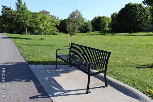 Stand-alone black metal bench in a park on a walkway surrounded with green grass lawn and trees on a sunny Summer day