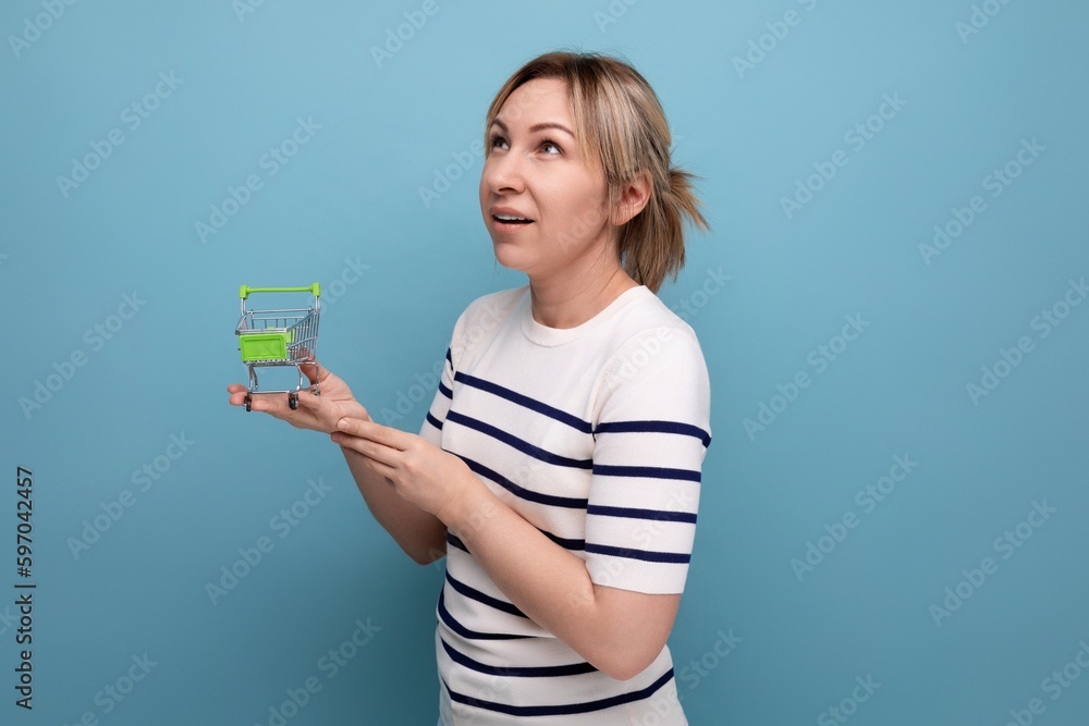 cute blond young mom shopaholic holding shopping cart and thinking how to spend money on blue background with copy space cute blond young mom shopaholic holding shopping cart and thinking how to spend money on blue background with copy space