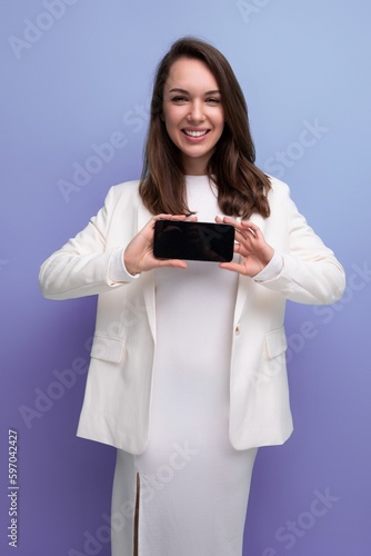 positive energetic young brunette with long hair woman in white dress holding smartphone with mockup