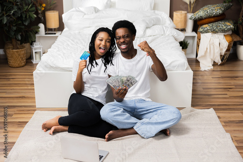 Fotografía Delighted happy lotery winners African American black couple sitting on floor at home smiling showing credit card and a lot of money in hands raising fists celebrating