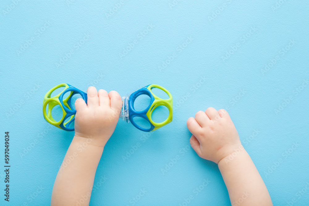 Baby boy hands playing with colorful rattle dumbbell on light blue table background. Pastel color. Closeup. Point of view shot. Toy of development for infant. Top down view. - obrazy, fototapety, plakaty 