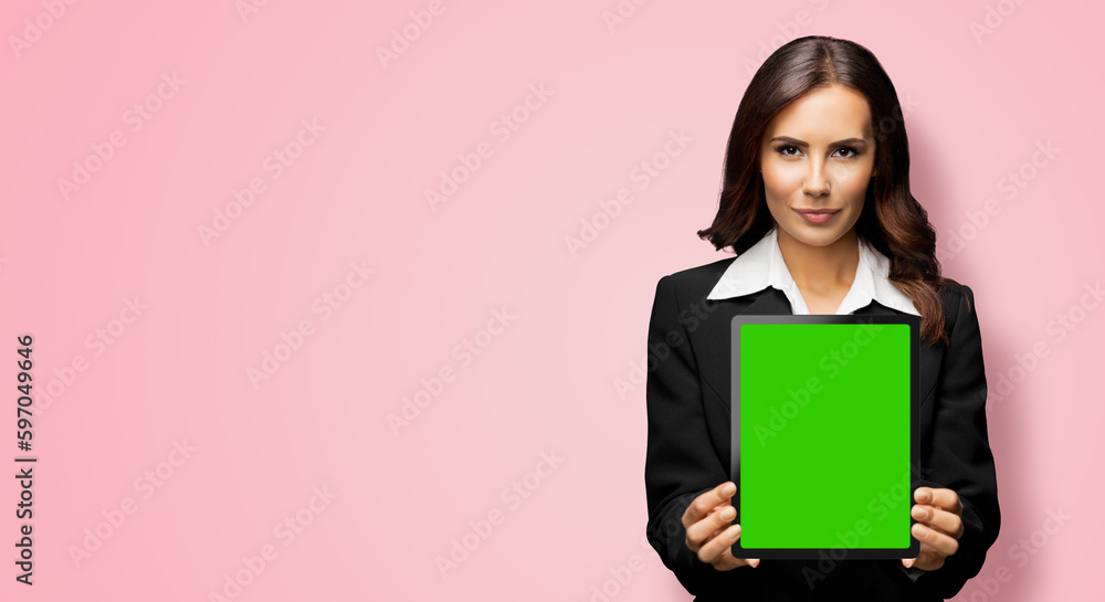 Obraz premium Portrait of serious brunette businesswoman showing tablet pc, touchpad, with green chroma key screen, on rose pink background. Confident attractive business woman at office.