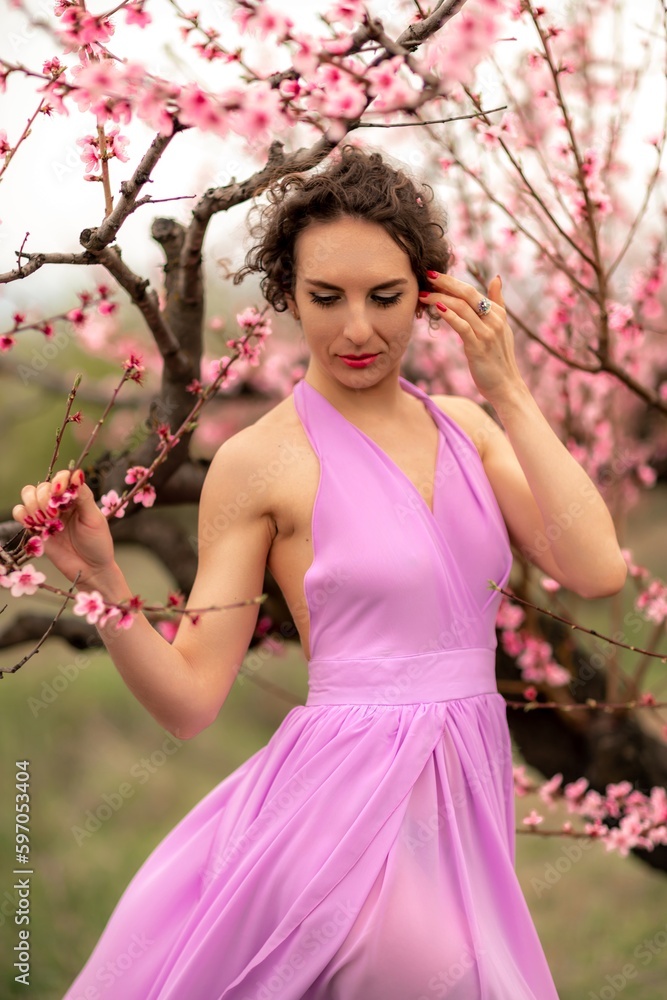Woman peach blossom. Happy curly woman in pink dress walking in the garden of blossoming peach trees in spring Woman peach blossom. Happy curly woman in pink dress walking in the garden of blossoming peach trees in spring