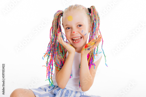 Portrait of delighted schoolgirl with colorful adorable stylish pigtails looking at camera smiling having hands full of colorful paints holding near face sitting on white background isolated.