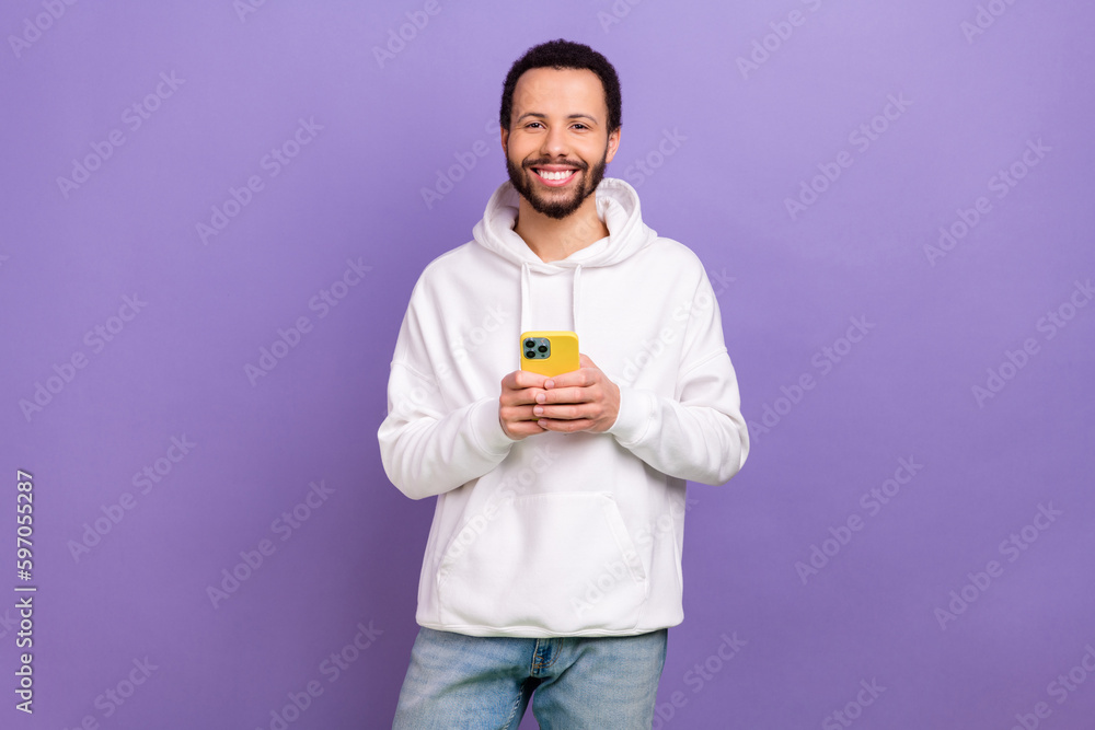 Photo of good mood satisfied man with curly hairstyle dressed white hoodie hold smartphone read email isolated on purple color background Photo of good mood satisfied man with curly hairstyle dressed white hoodie hold smartphone read email isolated on purple color background