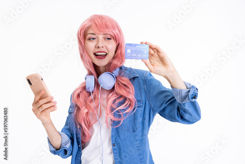 Smiled young woman holding credit card and smartphone in hands showing at camera wearing pink wig and modern headphones on neck posing for camera advertisiment of bank. photo