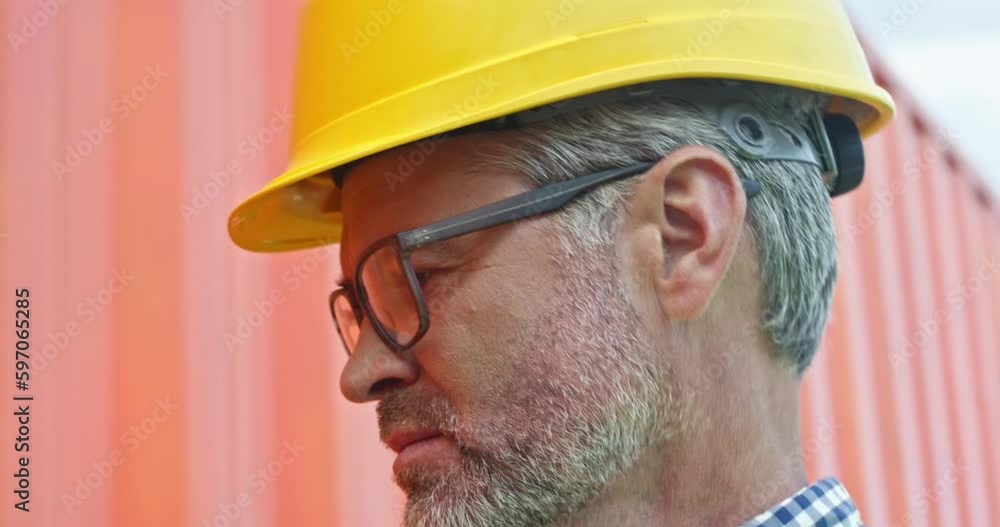 Close up of handsome Caucasian worker with grey hair and beard wearing glasses and yellow helmet. Standing near red container and considering something. Concept of safety equipment. Close up of handsome Caucasian worker with grey hair and beard wearing glasses and yellow helmet. Standing near red container and considering something. Concept of safety equipment.