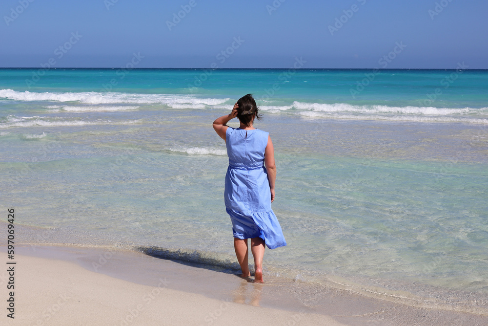 Barefoot woman in blue dress walking by sand against the azure waves. Travel and beach vacation on ocean coast Barefoot woman in blue dress walking by sand against the azure waves. Travel and beach vacation on ocean coast