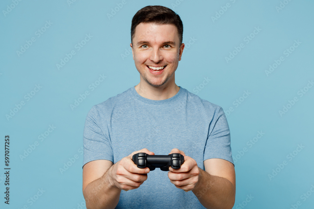 Young satisfied positive smiling happy man wear casual t-shirt hold in hand play pc game with joystick console isolated on plain pastel light blue cyan background studio portrait. Lifestyle concept. Young satisfied positive smiling happy man wear casual t-shirt hold in hand play pc game with joystick console isolated on plain pastel light blue cyan background studio portrait. Lifestyle concept.