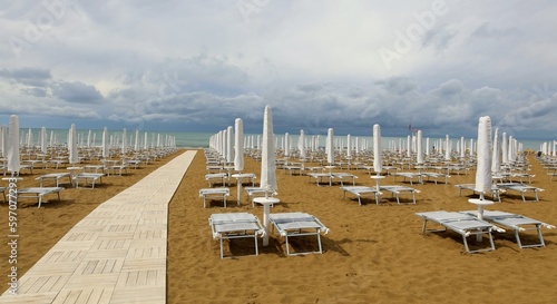 Empty beach resort under a dark cloudy sky. White beach umbrellas and tanning beds closed and wooden footpath without people. Background for copy space.