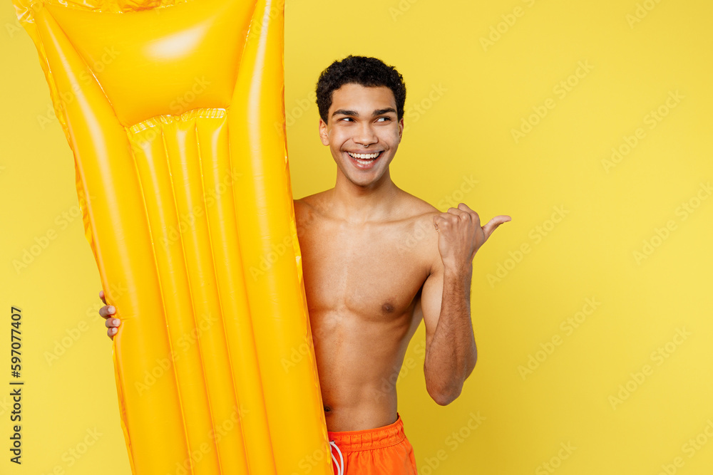 Young man wear orange shorts swimsuit relax near hotel pool hold in hand inflatable mattress point finger aside on area isolated on plain yellow background. Summer vacation sea rest sun tan concept. Young man wear orange shorts swimsuit relax near hotel pool hold in hand inflatable mattress point finger aside on area isolated on plain yellow background. Summer vacation sea rest sun tan concept.