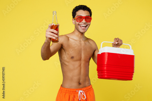 Foto Handsome young man wear orange shorts swimsuit glasses hold in hands beer bootle beach refrigerator relax near hotel pool isolated on plain yellow background