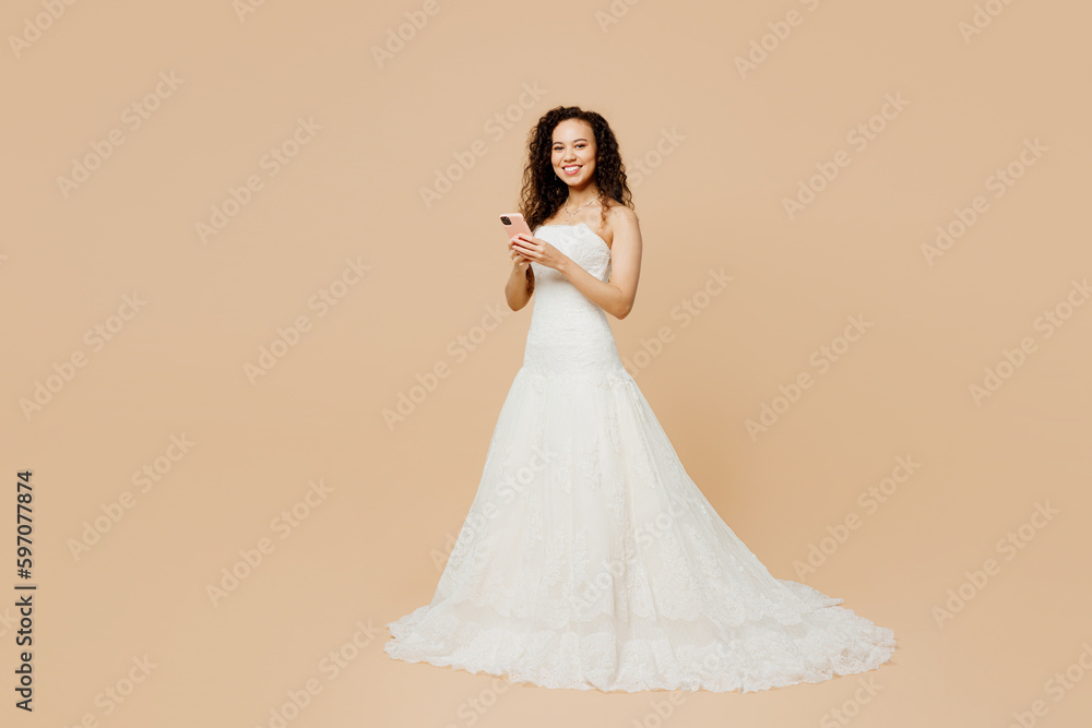 Full body fun happy young woman bride wear wedding dress posing hold in hand use mobile cell phone isolated on plain pastel light beige background studio portrait. Ceremony celebration party concept. Full body fun happy young woman bride wear wedding dress posing hold in hand use mobile cell phone isolated on plain pastel light beige background studio portrait. Ceremony celebration party concept.