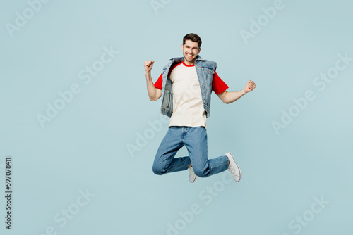 Full body young man wear denim vest red t-shirt casual clothes jump high do winner gesture celebrate clench fists say yes isolated on plain pastel light blue cyan background studio. Lifestyle concept.