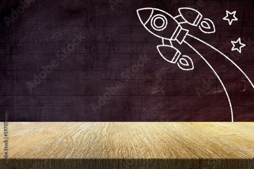 Fototapeta Naklejka Na Ścianę i Meble -  Wood Table with Retro Rocket in Chalk Drawing Style on Old Grunge Chalkboard Background, Suitable for Product Presentation Backdrop, Display, and Mock up in Back to School and Education Concept.