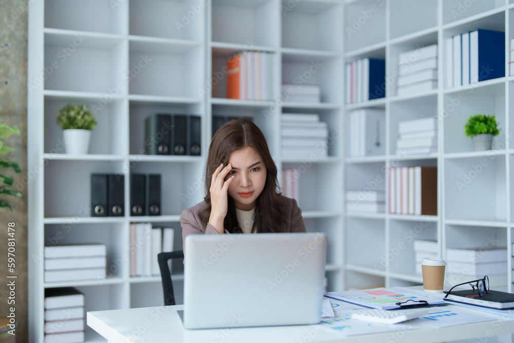 Portrait of a young Asian woman showing a serious face as she uses her calculator, financial documents and computer laptop on her desk in the early morning hours. Portrait of a young Asian woman showing a serious face as she uses her calculator, financial documents and computer laptop on her desk in the early morning hours.