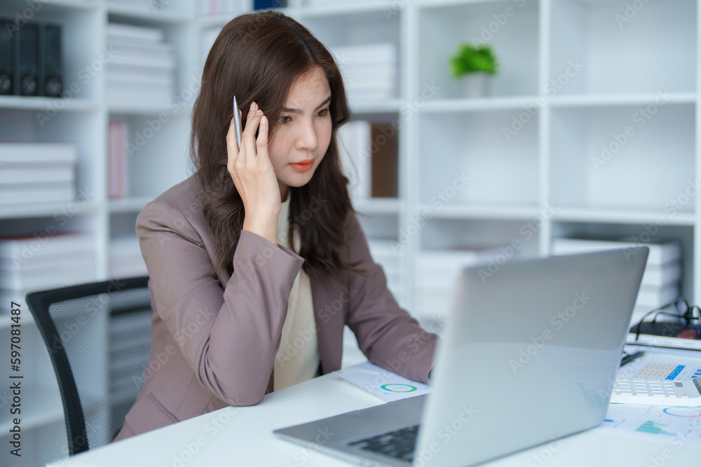 Portrait of a young Asian woman showing a serious face as she uses her calculator, financial documents and computer laptop on her desk in the early morning hours. Portrait of a young Asian woman showing a serious face as she uses her calculator, financial documents and computer laptop on her desk in the early morning hours.