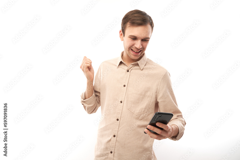 Portrait of young handsome caucasian man using mobile phone and smiling, clenching fist celebrating victory isolated on white studio background, joyful facial expression Portrait of young handsome caucasian man using mobile phone and smiling, clenching fist celebrating victory isolated on white studio background, joyful facial expression