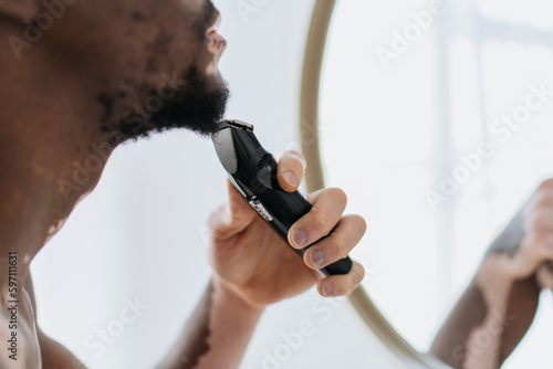 Cropped view of blurred african american man with vitiligo shaving with electric razor in bathroom.