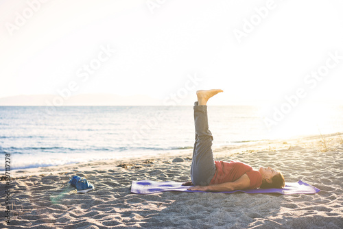 Senior woman exercising, stretching on yoga mat at the beach, doing leg raises. © belekekin