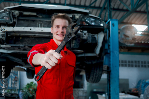 Hand holding spanner with blurred background of mechanic man in red uniform work with lifted vehicle, auto mechanic checking repairing and maintenance customer car automobile at garage service shop.