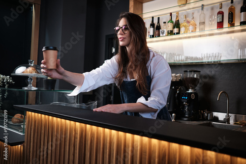 A friendly barista giving coffee to go to a customer while standing behind a bar counter.