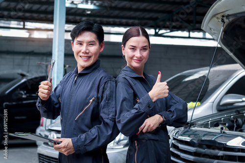 Portrait of smiling Asian mechanic man and beautiful woman in uniform standing and give thump up at garage vehicle shop, auto mechanic technician maintenance customer car automobile at repair service.
