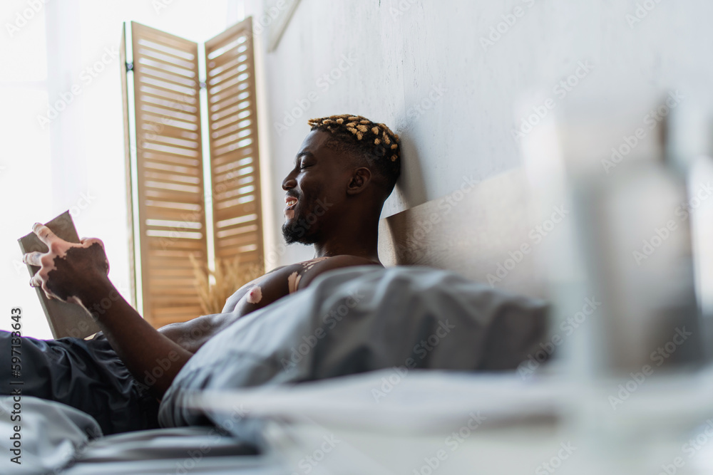 Side view of cheerful and shirtless african american man with vitiligo reading book on bed. Side view of cheerful and shirtless african american man with vitiligo reading book on bed.