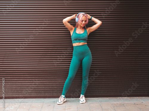 Fitness smiling woman in green sports clothing with pink hair. Young beautiful model with perfect body.Female in the street near roller shutter wall.Listening music in wireless headphones