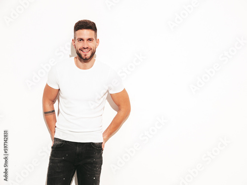 Portrait of handsome smiling stylish hipster lambersexual model. Man dressed in white T-shirt and jeans. Fashion male posing in studio