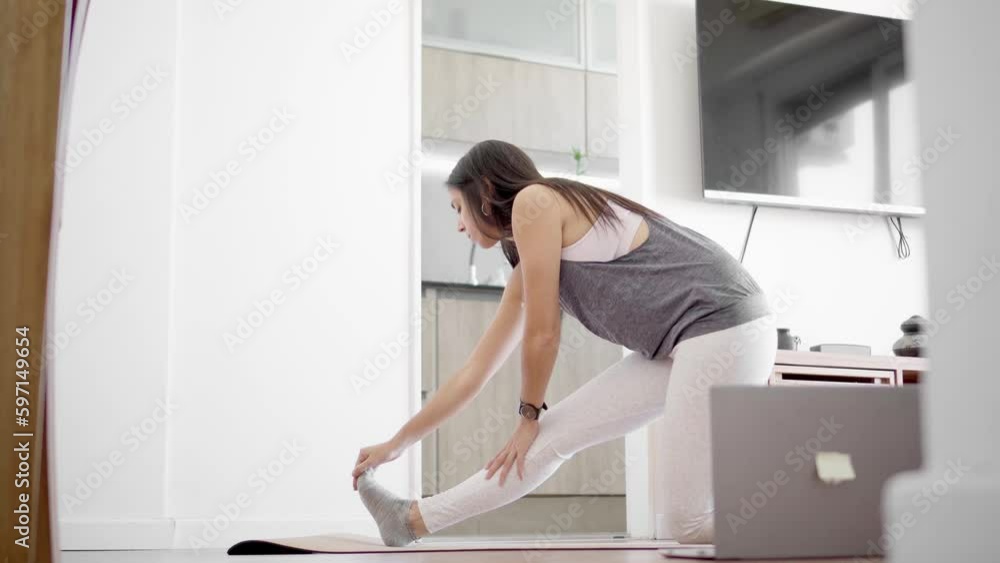 Young latin woman in sportswear doing leg stretches in her living room with her laptop open. Young latin woman in sportswear doing leg stretches in her living room with her laptop open.