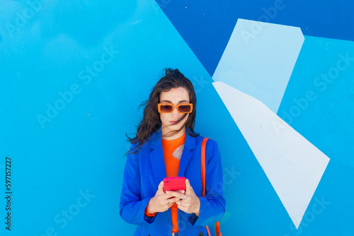 Fashionable young woman using smart phone in front of blue wall