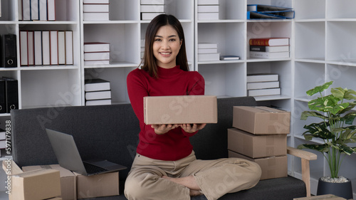 Portrait of young pretty small business owner handing the box to the camera while sitting in a room full of boxes. © PaeGAG