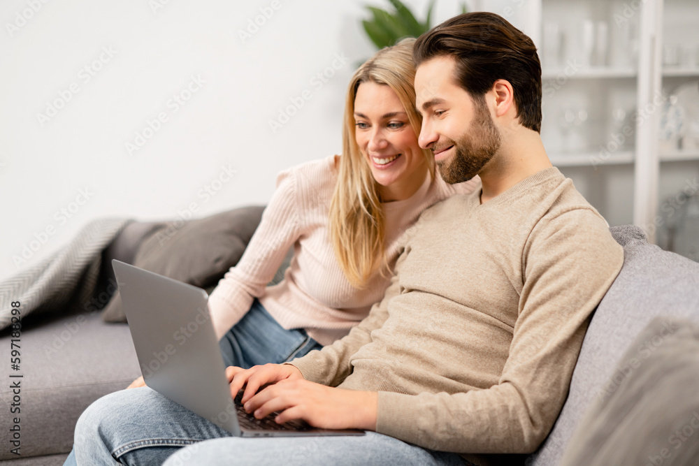 Overjoyed couple in love spending leisure time online with laptop at home. Blonde woman and man looking at the laptop screen while sitting on the sofa and watching comedy movies Overjoyed couple in love spending leisure time online with laptop at home. Blonde woman and man looking at the laptop screen while sitting on the sofa and watching comedy movies