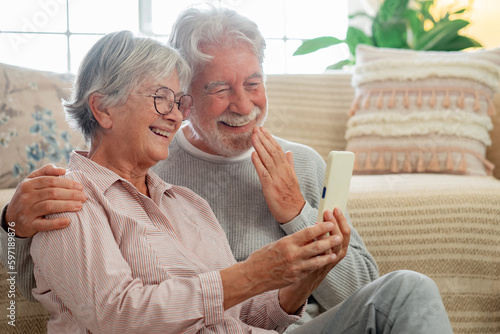 Video call concept. Smiling senior family couple sitting on the floor at home in video connection by smartphone romantic elderly man and woman getting closer enjoying moment of affection cuddling