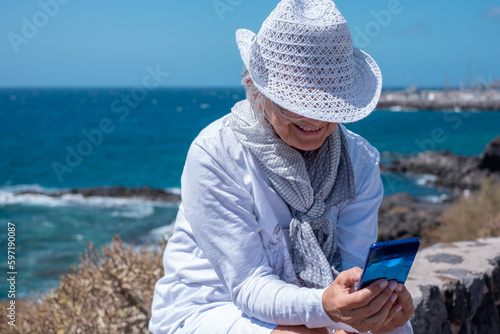 Happy Senior Mature Woman Sitting Outdoor Feeling in Vacation at Sea. Elderly Female Wearing Hat Enjoying Freedom and Relax using Smartphone