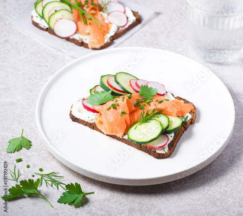 Smorrebrod, toasted bread with rye bread, salmon,curd cheese with herbs, green wild onion, cucumber and radish on the white plate. Hearty and healthy Scandinavian snack, lunch. Summer menu
