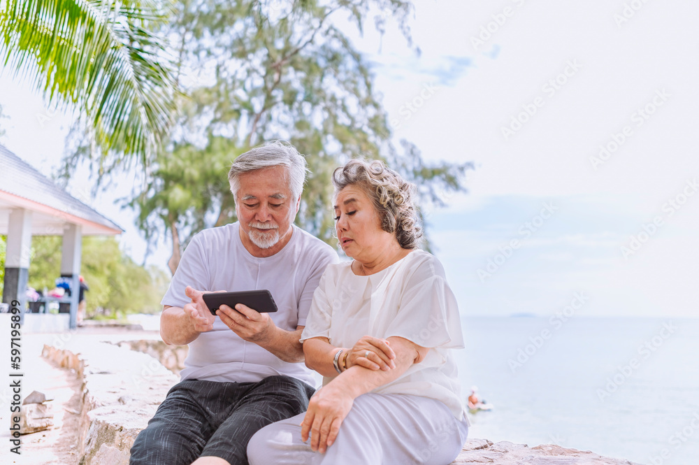 Happy asian senior retired couple; relax elder man and woman enjoying with smartphone apps enjoy online entertainments in retired vacation at sea beach outdoor. Family outdoor lifestyle