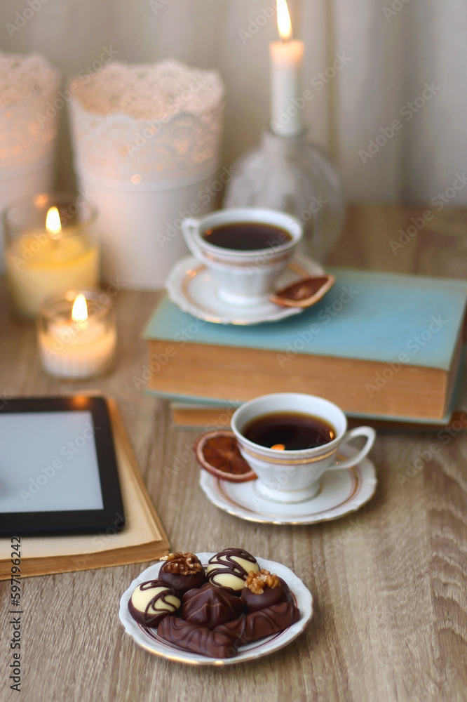 Books, reading glasses, e-reader, plate of chocolate pralines, bowl of cookies, cups of tea and lit candles on the table. Selective focus.