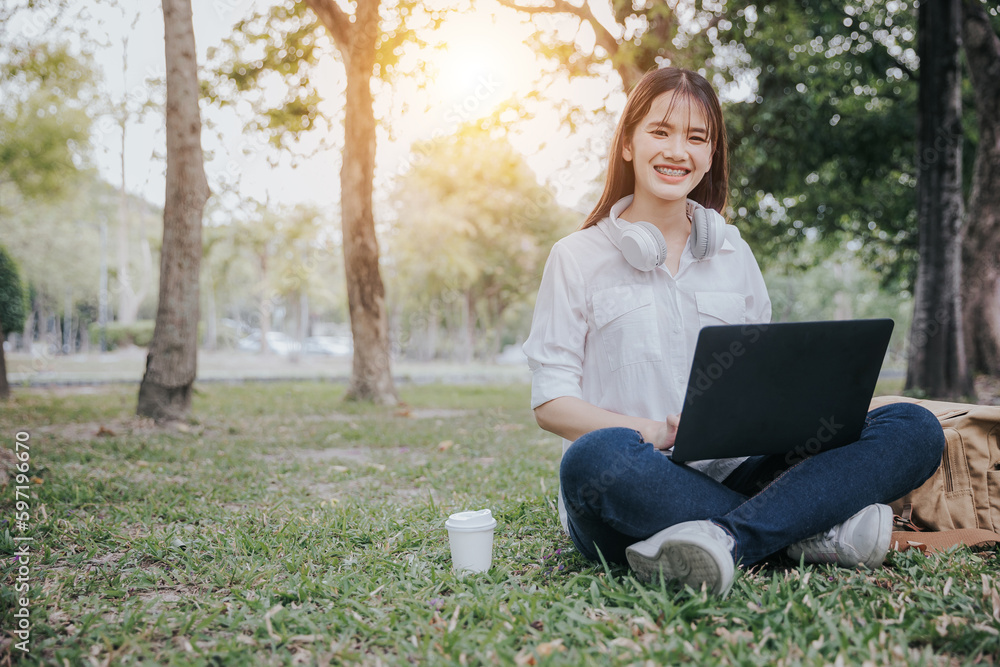 Portrait of young asian woman in white shirt sitting on green grass in park with legs crossed during summer day while using laptop and wireless headphones for video call.