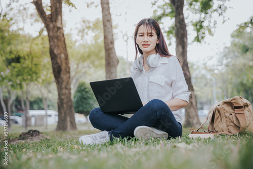 Portrait of young asian woman in white shirt sitting on green grass in park with legs crossed during summer day while using laptop and wireless headphones for video call.