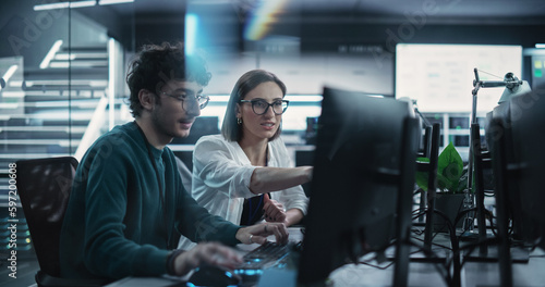 Two Young Colleagues Working on Computers and Talking at a Workplace. Female and Male Software Developers Discussing a Solution for Their Collaborative Artificial Intelligence Project photo